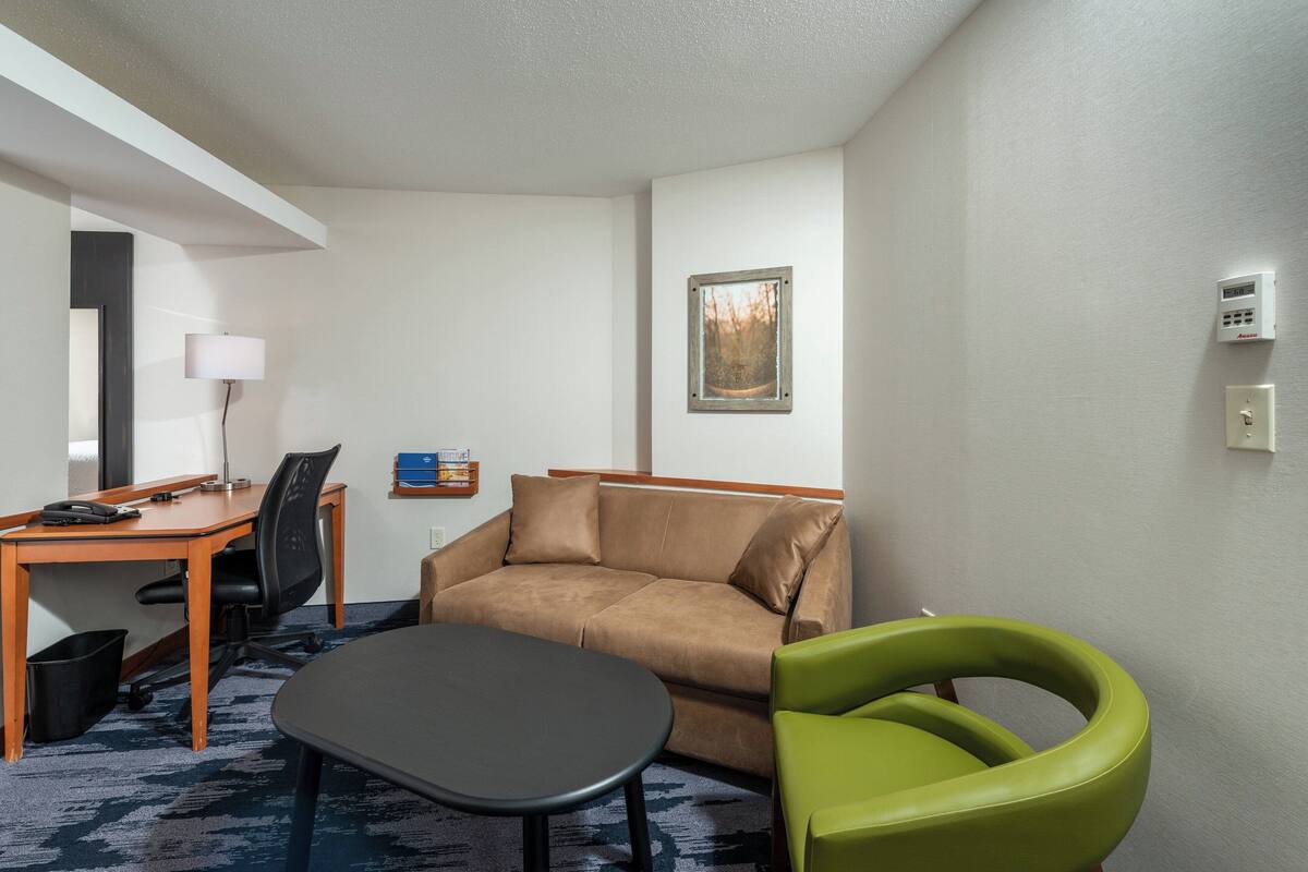 Una habitación de hotel con un sofá marrón, una silla verde, una mesita pequeña, un escritorio con una lámpara, un cuadro en la pared y un termostato.