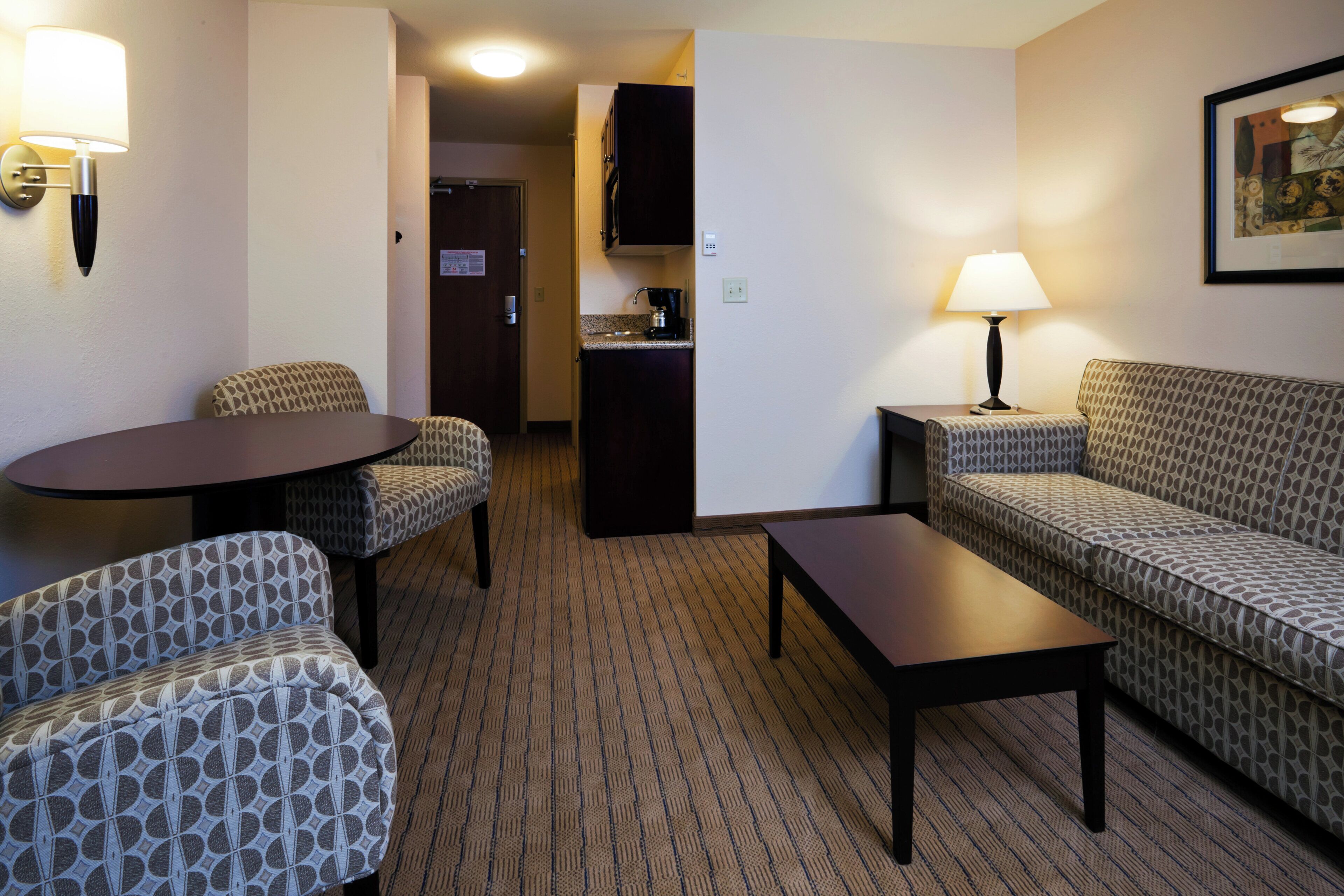 A hotel room with a sofa, two armchairs, a small table, a lamp, a kitchen area, and a framed picture on the wall.