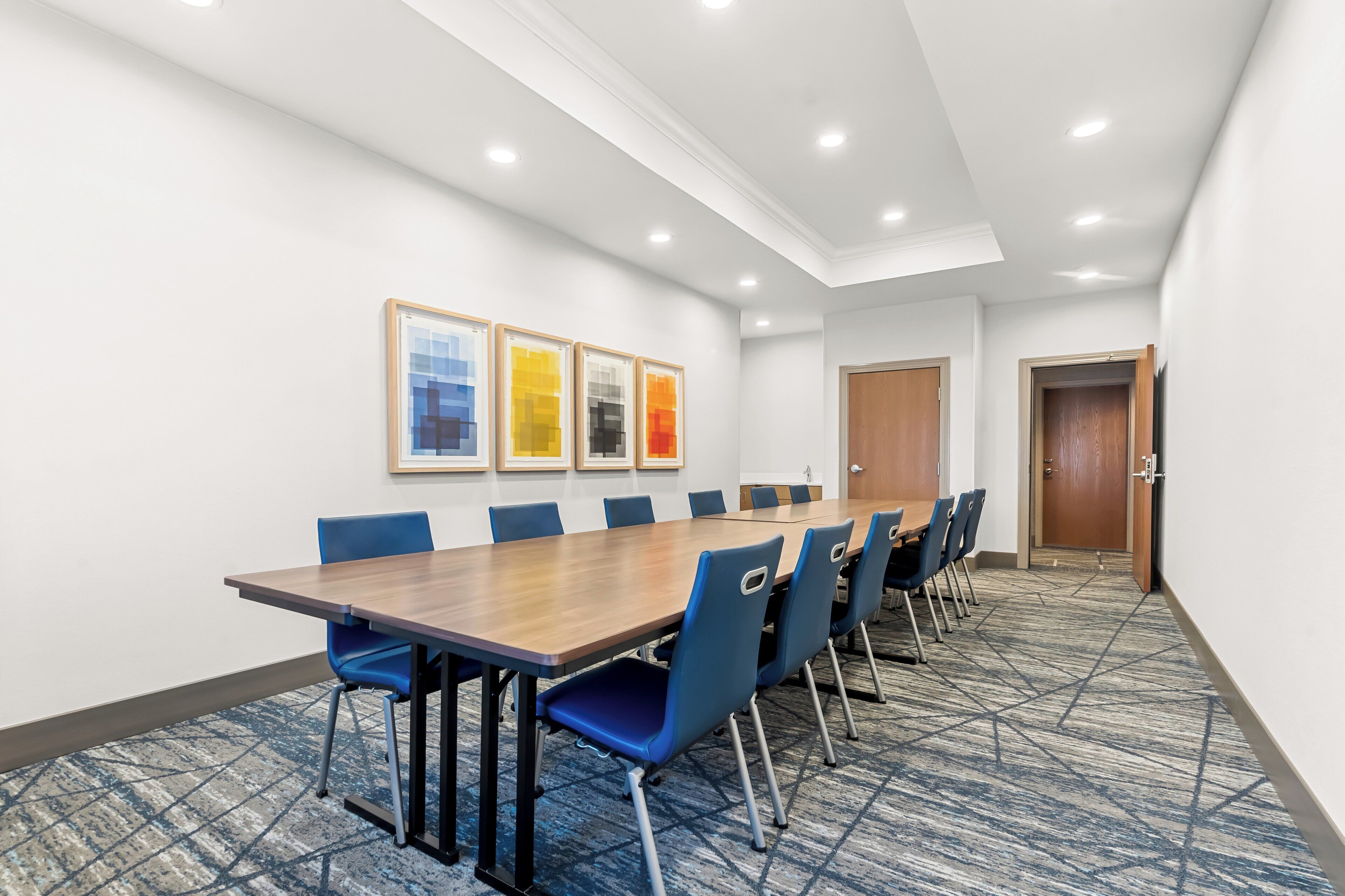 A conference room with a long table, blue chairs, framed artwork on the wall, and a carpeted floor.
