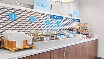 Zona de desayuno tipo buffet con estaciones de comida señalizadas, que incluyen huevos revueltos, tocino y salchicha.