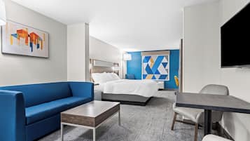 Une chambre d’hôtel avec un canapé bleu, une table basse en bois, un lit avec une literie blanche, une télévision et une œuvre d’art géométrique accrochée au mur.