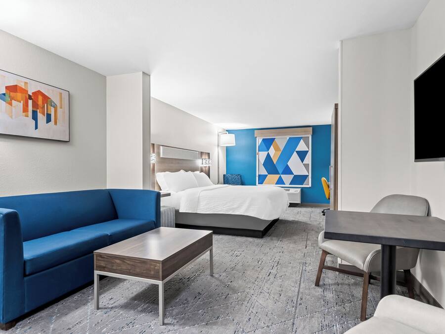 Une chambre d’hôtel avec un canapé bleu, une table basse en bois, un lit avec une literie blanche, une télévision et une œuvre d’art géométrique accrochée au mur.