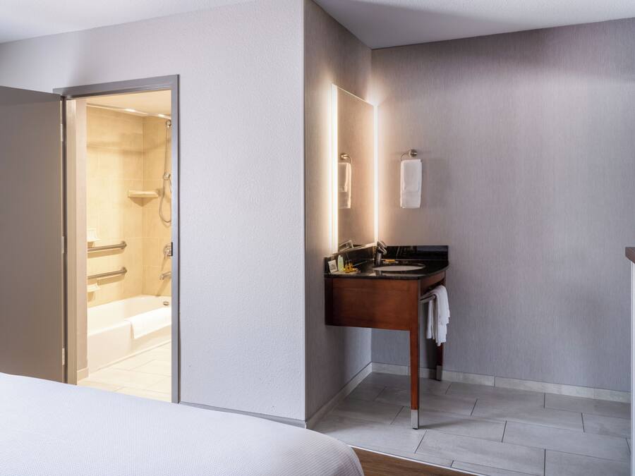 Una habitación de hotel moderna con suelo de madera, una cama, un baño con ducha, lavamanos y espejo.