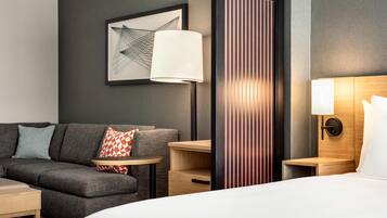 A hotel room with a bed, a sofa, a small table, a lamp, a nightstand, and a wall-mounted artwork.