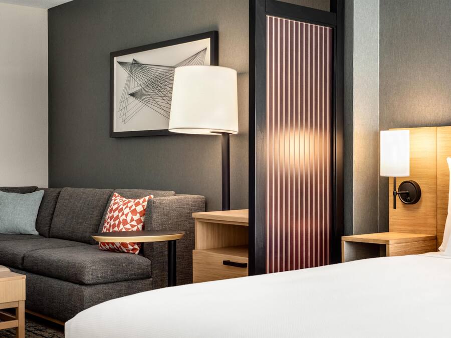Habitación de hotel con cama, sofá, una mesita, lámpara, mesita de noche y una obra de arte colgada en la pared.