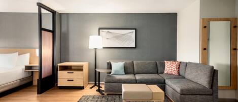 A modern hotel room with a bed, a grey sofa, a small table, a lamp, and a framed picture on the wall.