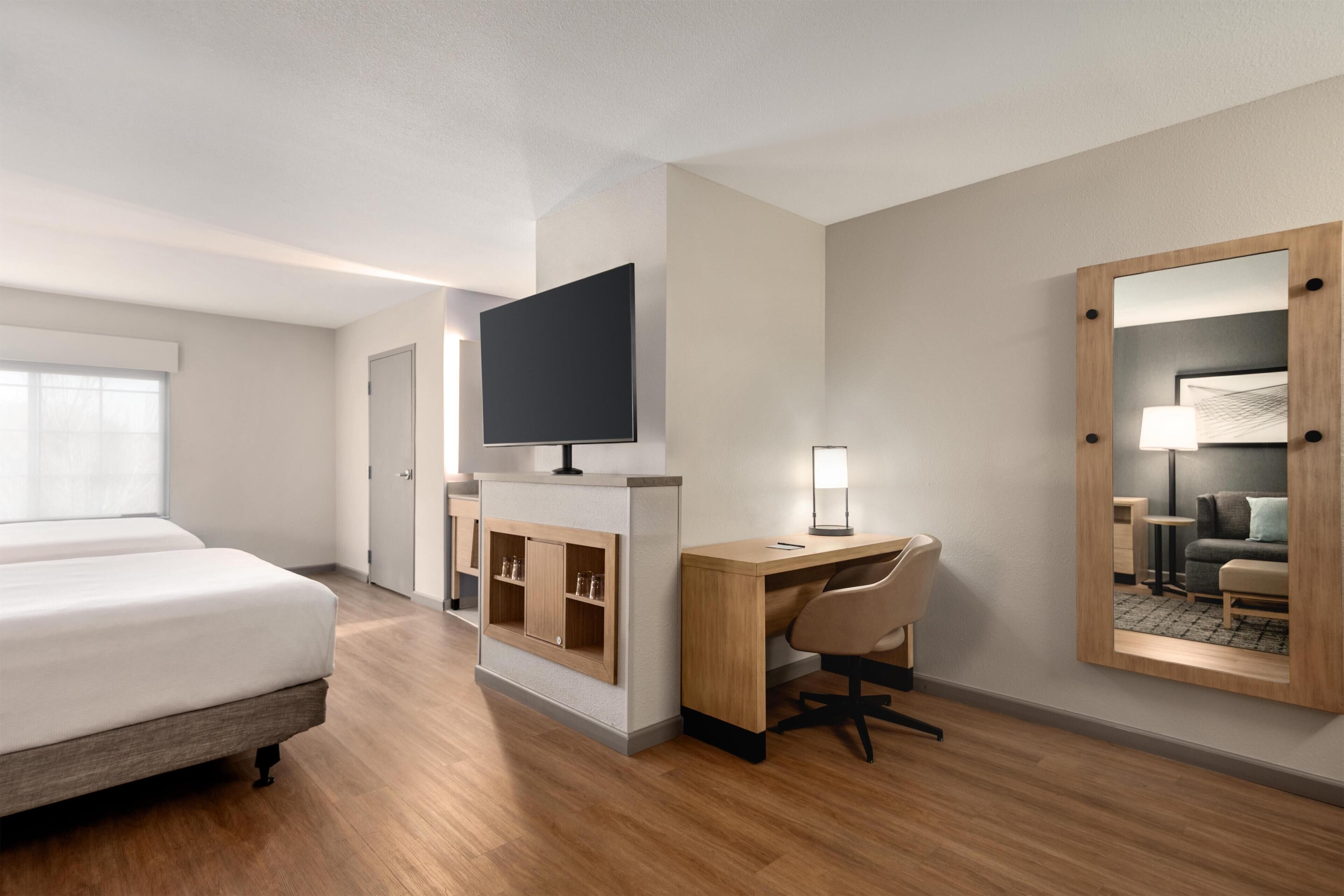 Una habitación de hotel con cama, escritorio, silla, televisor y espejo.
