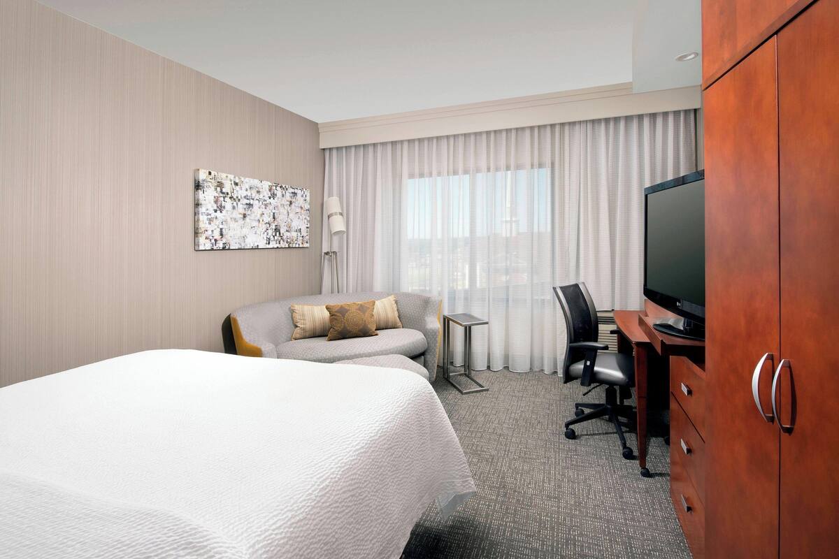 Habitación de hotel con una cama grande, un sofá, un escritorio con computadora, televisión y un ventanal con cortinas.