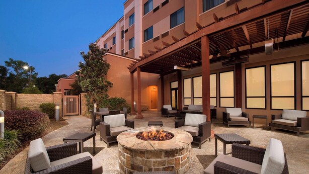 An outdoor patio with a stone fire pit surrounded by wicker furniture, a covered seating area, and a building with large windows in the background. - Courtyard by Marriott Tyler (Tyler)