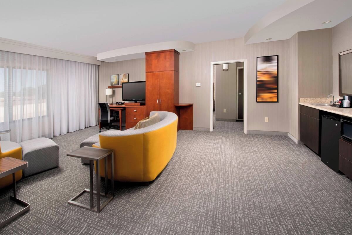 Habitación de hotel con sala de estar, kitchenette, televisor de pantalla plana y un gran ventanal con cortinas.
