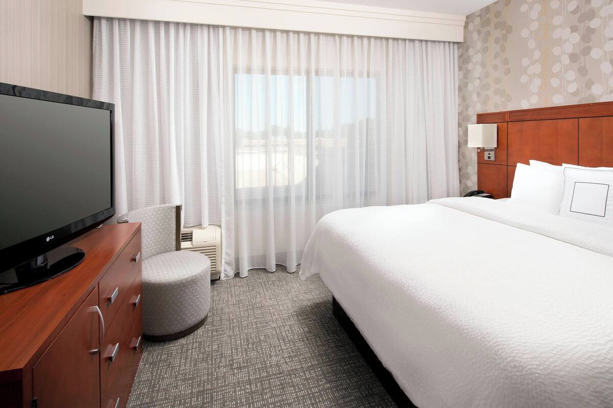 Una habitación de hotel con una cama grande, un televisor sobre un mueble de madera, una silla redonda pequeña y una mesita de noche con una lámpara.