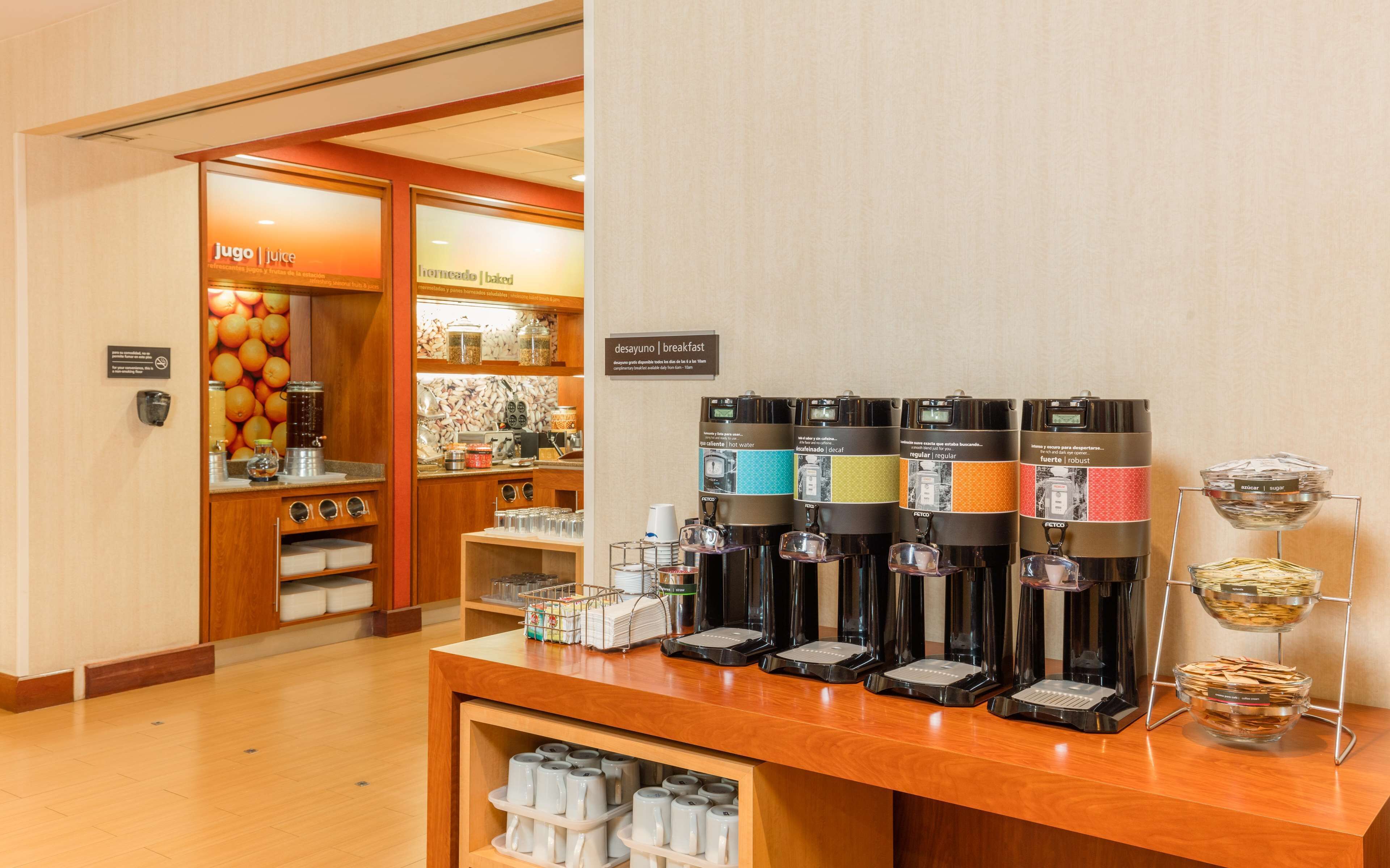 Un área de desayuno con varias máquinas de café, un dispensador de zumo y una presentación de productos para el desayuno.