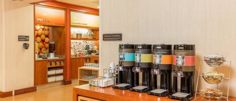 A breakfast area with multiple coffee machines, a juice dispenser, and a display of breakfast items.