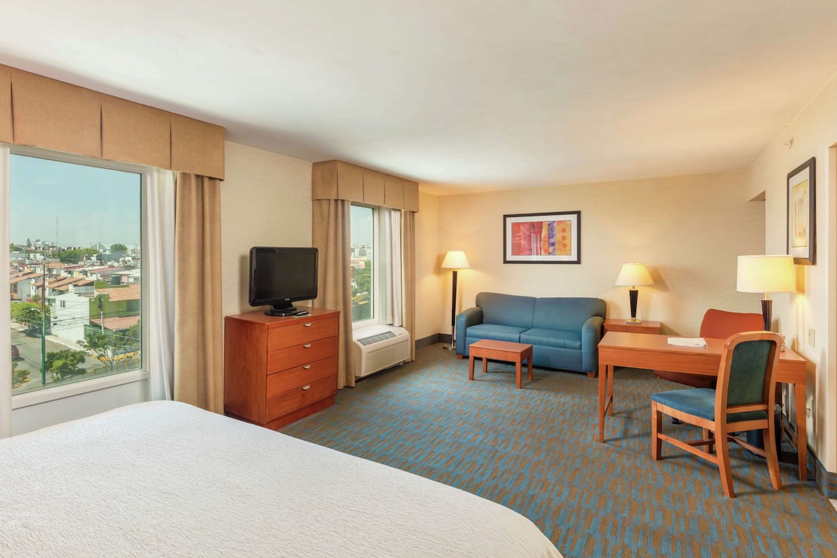 A hotel room with a bed, a desk with a chair, a sofa, a television, and a large window offering a city view.