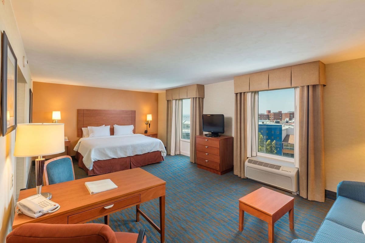 A hotel room with a bed, desk, chair, TV, and a view of the city.