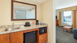 Quarto de hotel com cozinha compacta, incluindo pia, fogão, micro-ondas e máquina de café. Há um espelho grande acima da pia e uma janela com vista para a cidade.