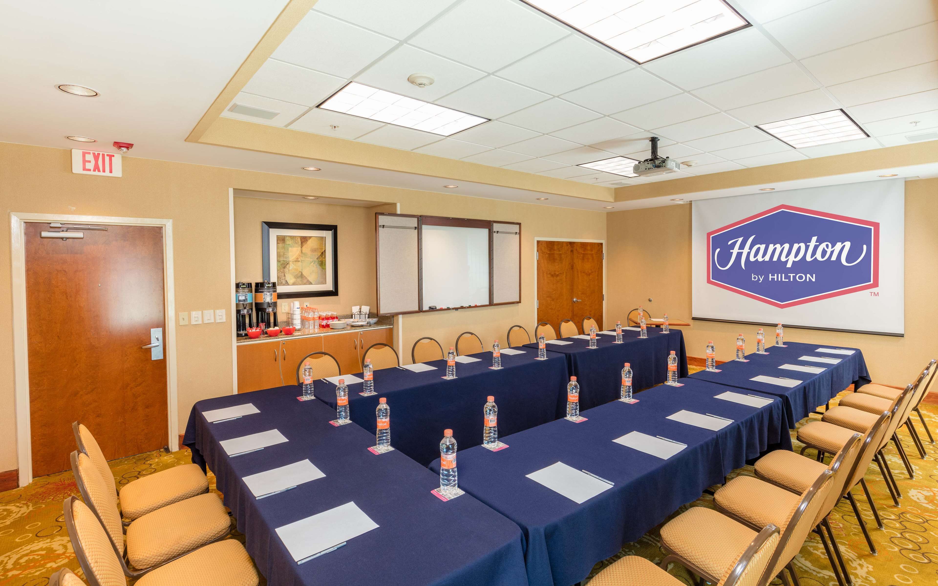 A conference room with a long table, chairs, a projector screen, and a sign reading "Hampton by Hilton".