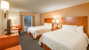 Quarto de hotel com duas camas, uma escrivaninha com computador, uma cadeira e vista para a paisagem urbana.