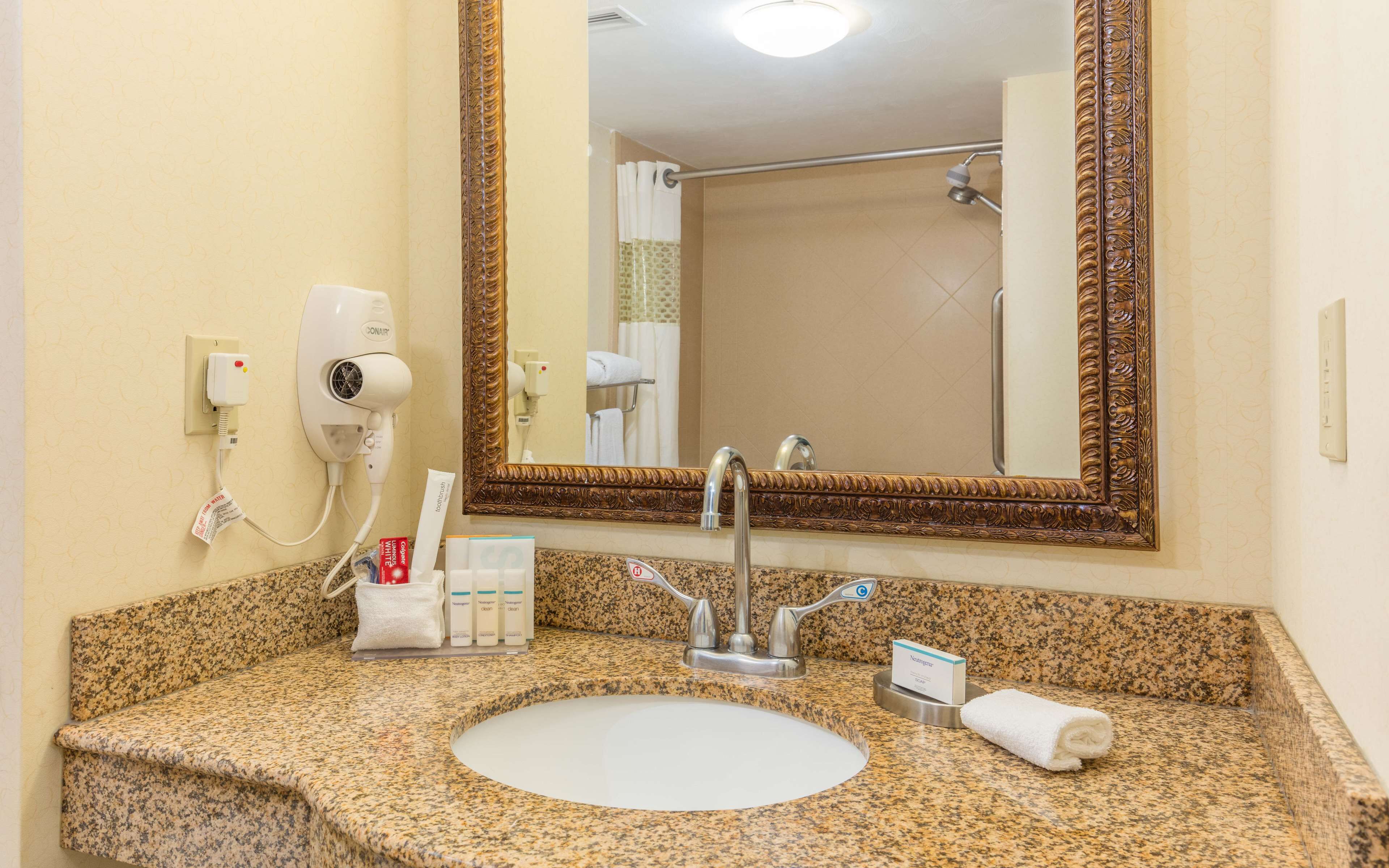 A bathroom with a granite countertop, a white sink, a large mirror with an ornate frame, a hairdryer, and toiletries.