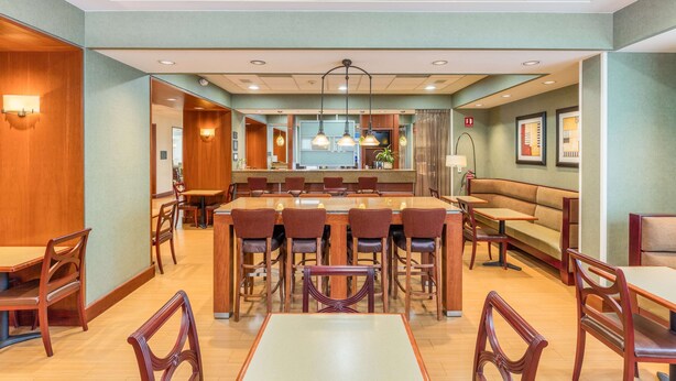 Uma sala de jantar de hotel com bar central, diversas opções de assento incluindo mesas e cabines, e um design de teto moderno.