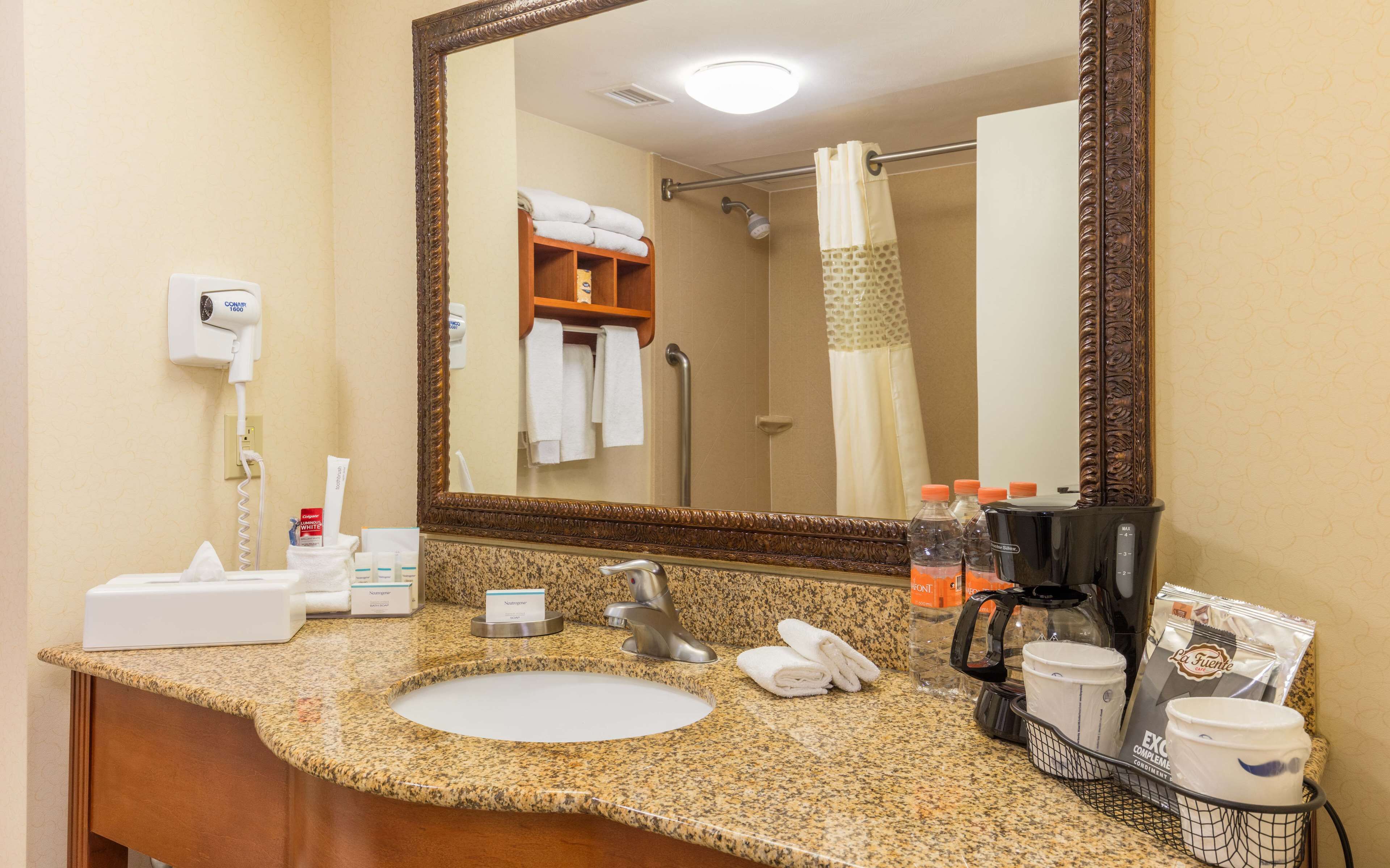 A hotel bathroom with a granite countertop sink, a large mirror, a hairdryer, a towel rack, and toiletries.