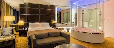 A modern hotel room with a large bed, a sitting area with a sofa and armchair, a separate bathroom with a glass-enclosed jacuzzi, and a large mirror.