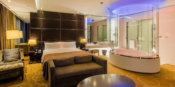 A modern hotel room with a large bed, a sitting area with a sofa and armchair, a separate bathroom with a glass-enclosed jacuzzi, and a large mirror.