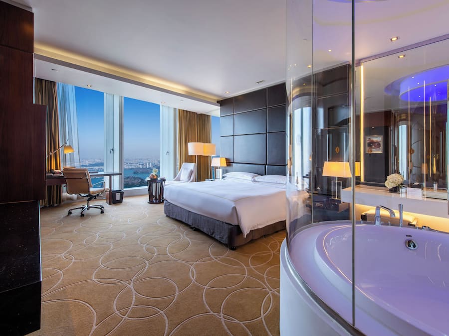 A hotel room with a large bed, a sitting area with a chair, a desk, and a view of the city.