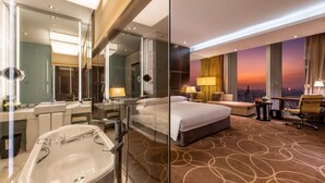Une chambre d’hôtel moderne avec un grand lit, une salle de bain séparée avec baignoire, un canapé et une vue sur la ville au coucher du soleil.