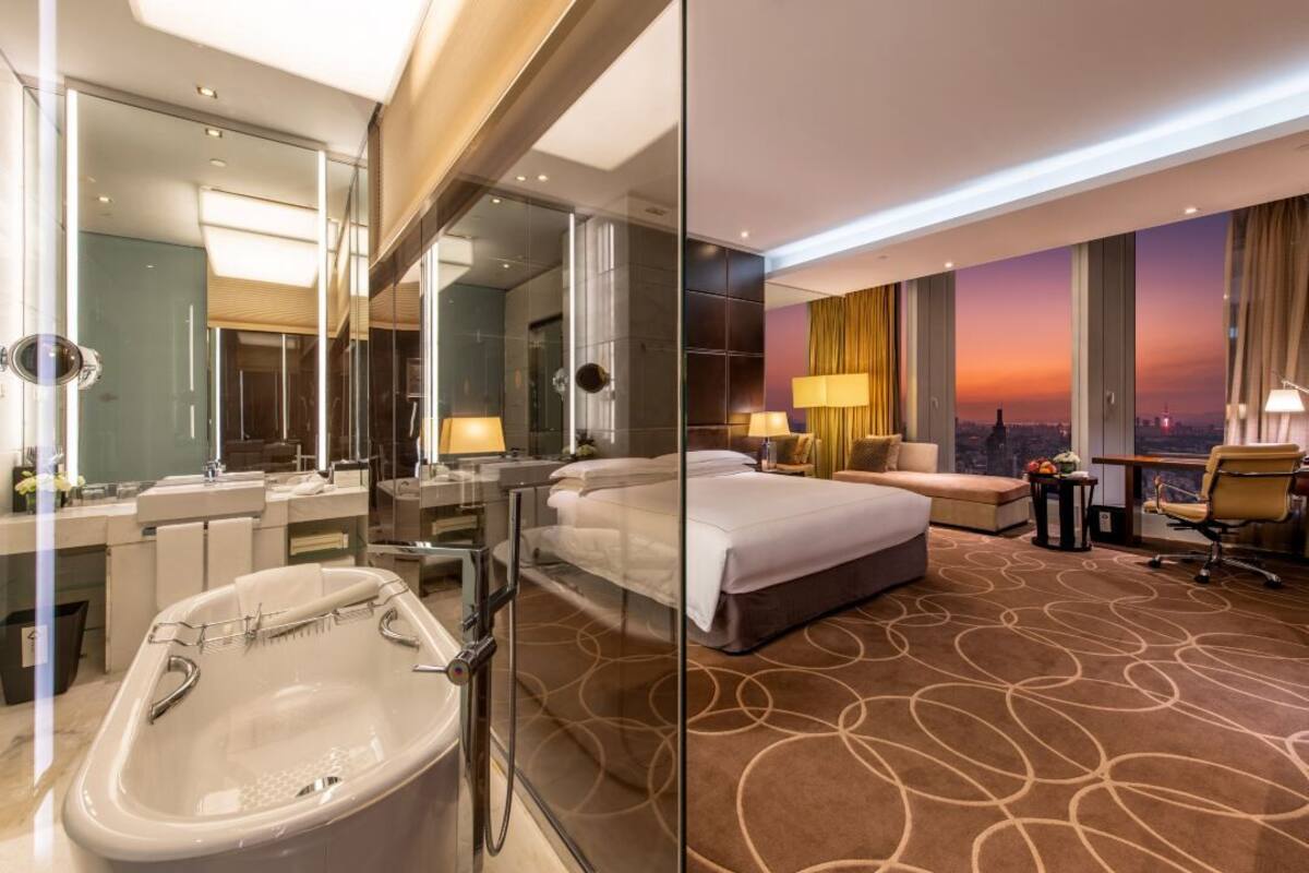 Quarto de hotel moderno com uma cama grande, banheiro separado com banheira, um sofá e vista para a cidade ao pôr do sol.