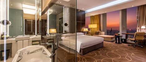 A modern hotel room with a large bed, a separate bathroom with a bathtub, a sofa, and a view of the city at sunset.