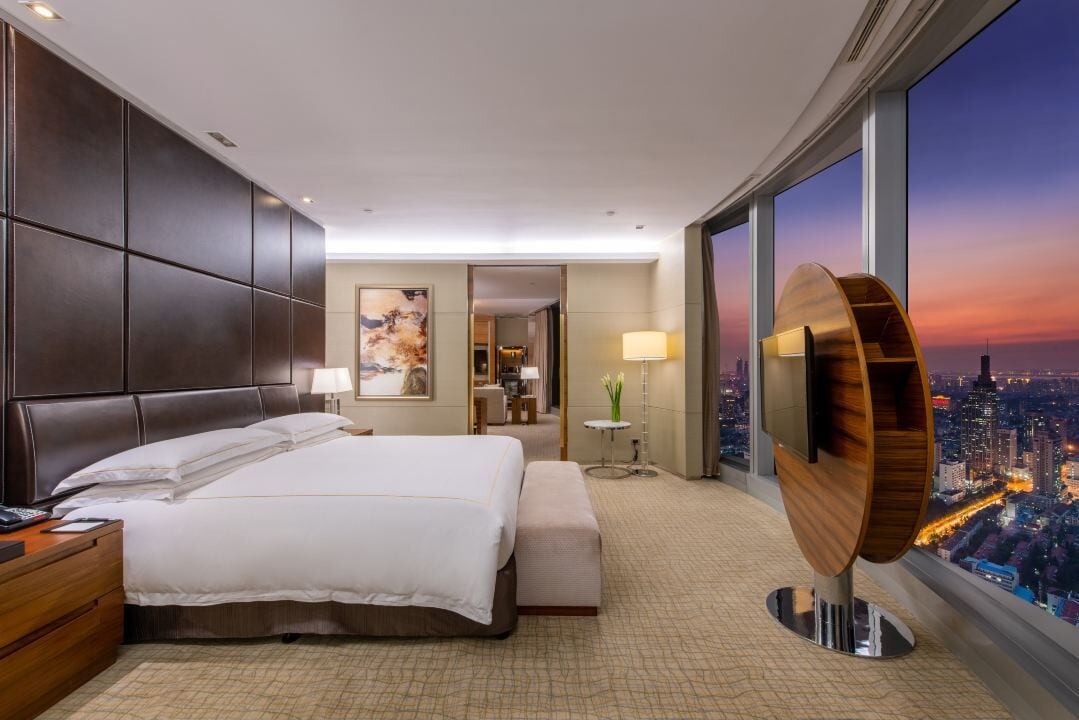 Ein modernes Hotelzimmer mit einem großen Bett, einem Schreibtisch und Blick auf die Stadt bei Sonnenuntergang.