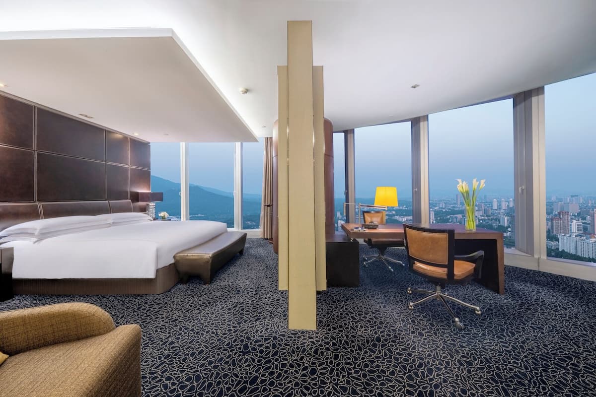Quarto de hotel com cama grande, escrivaninha com cadeira e vista para a paisagem urbana, além de uma janela ampla.