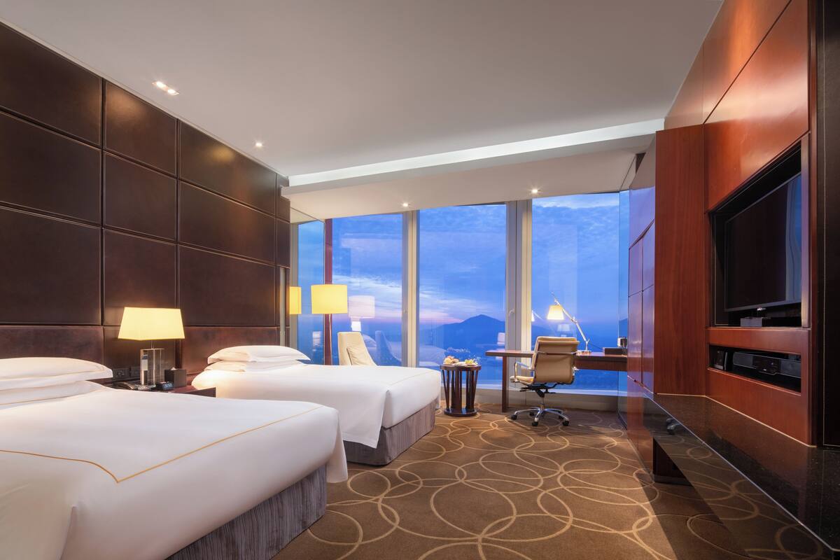 Quarto de hotel com duas camas, uma escrivaninha, televisão e uma janela grande com vista para a cidade.