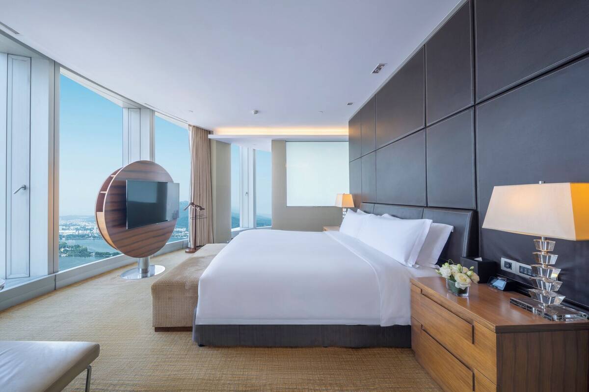 Quarto de hotel moderno com uma cama grande, televisão, mesa de cabeceira com abajur e vista para a cidade.