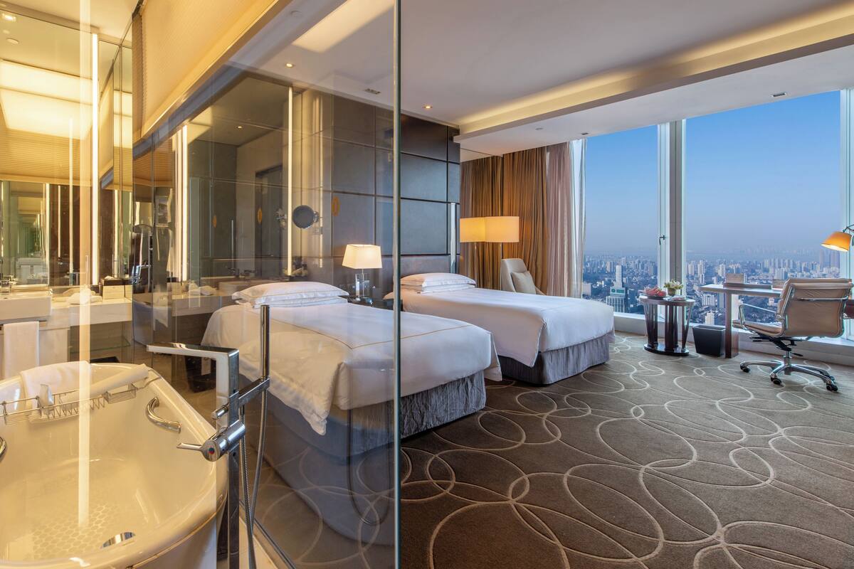 Quarto de hotel com uma banheira grande, duas camas, uma mesa de trabalho com cadeira e vista para a cidade.