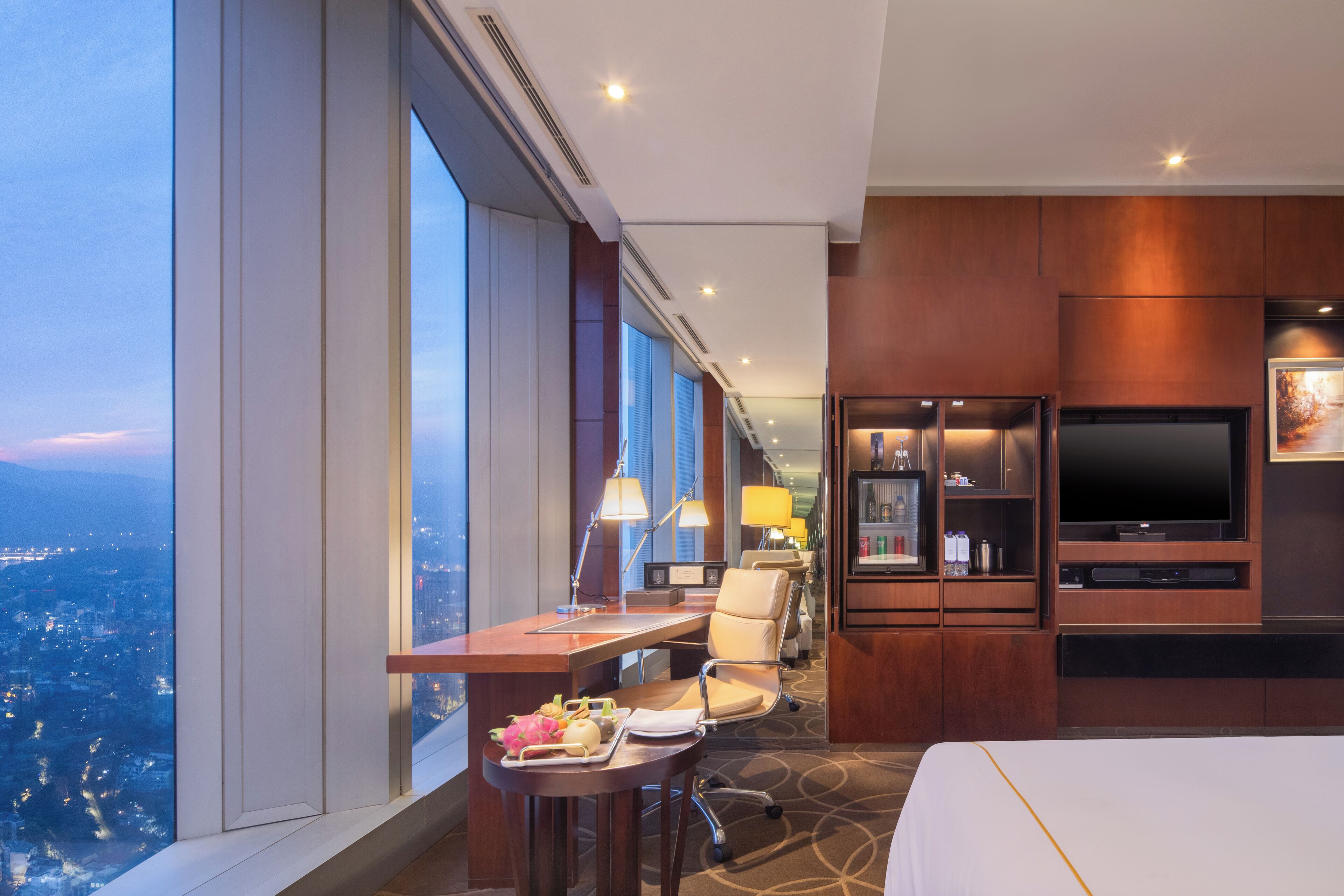 Ett hotellrum med ett stort fönster, ett skrivbord med en stol, en säng, en TV och en minibar.
