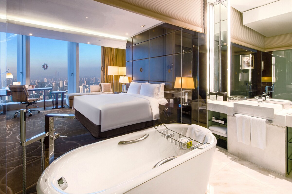 A modern hotel room with a large bed, a bathtub, a vanity with a mirror, and a city view.