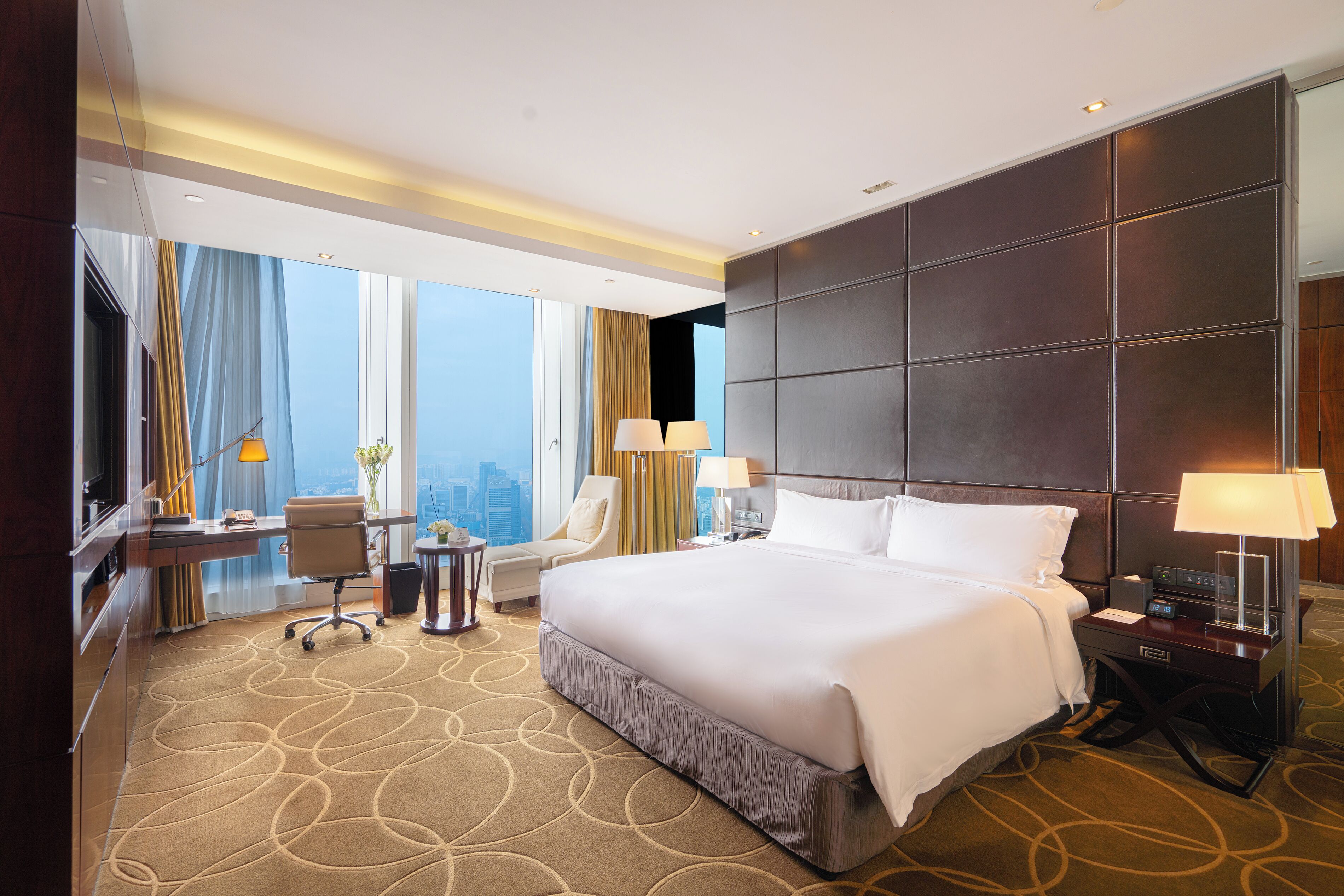 A modern hotel room with a large bed, a desk, a chair, and a view of the city.