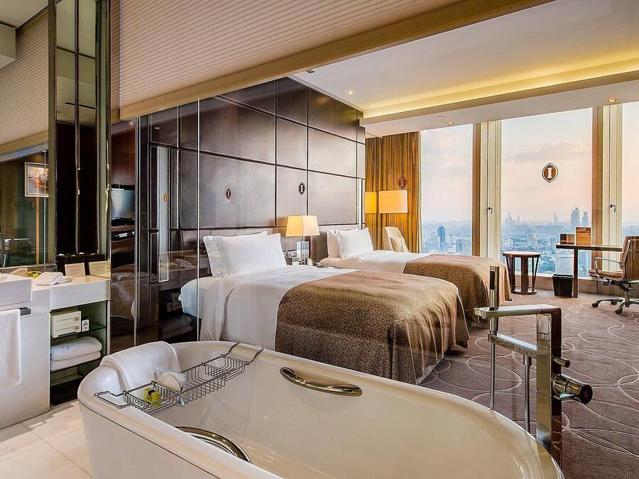 A hotel room with a large bathtub, two beds, a desk, and a view of the city.