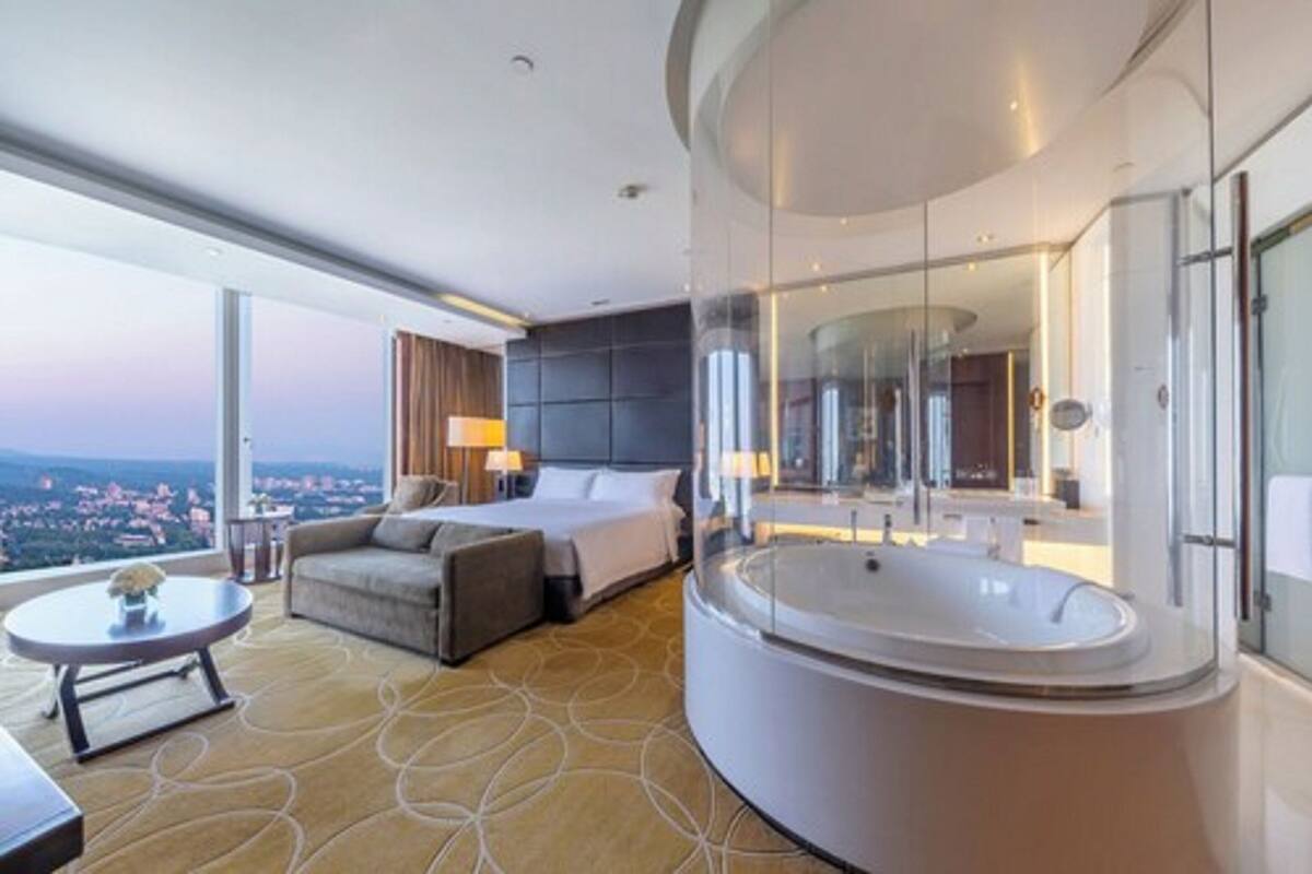 Quarto de hotel moderno com uma cama grande, área de estar, banheiro com paredes de vidro e vista para a cidade.