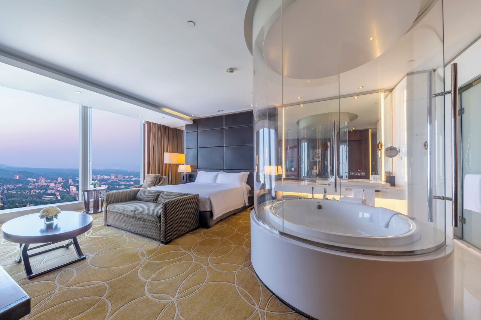 A modern hotel room with a large bed, a sitting area, a round bathtub, and a city view.