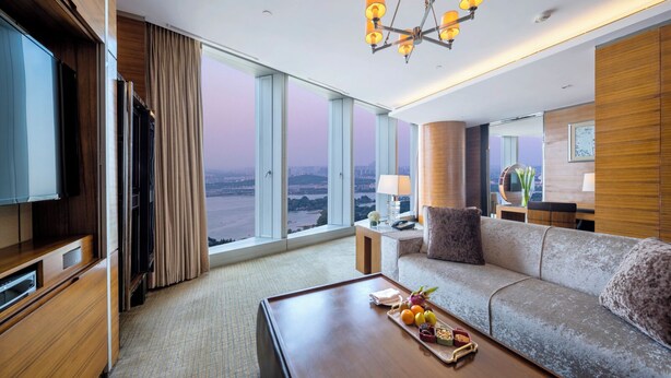 Une chambre d’hôtel moderne dotée d’une grande fenêtre, d’un téléviseur à écran plat, d’un canapé, d’une table basse avec des fruits et d’un lustre.