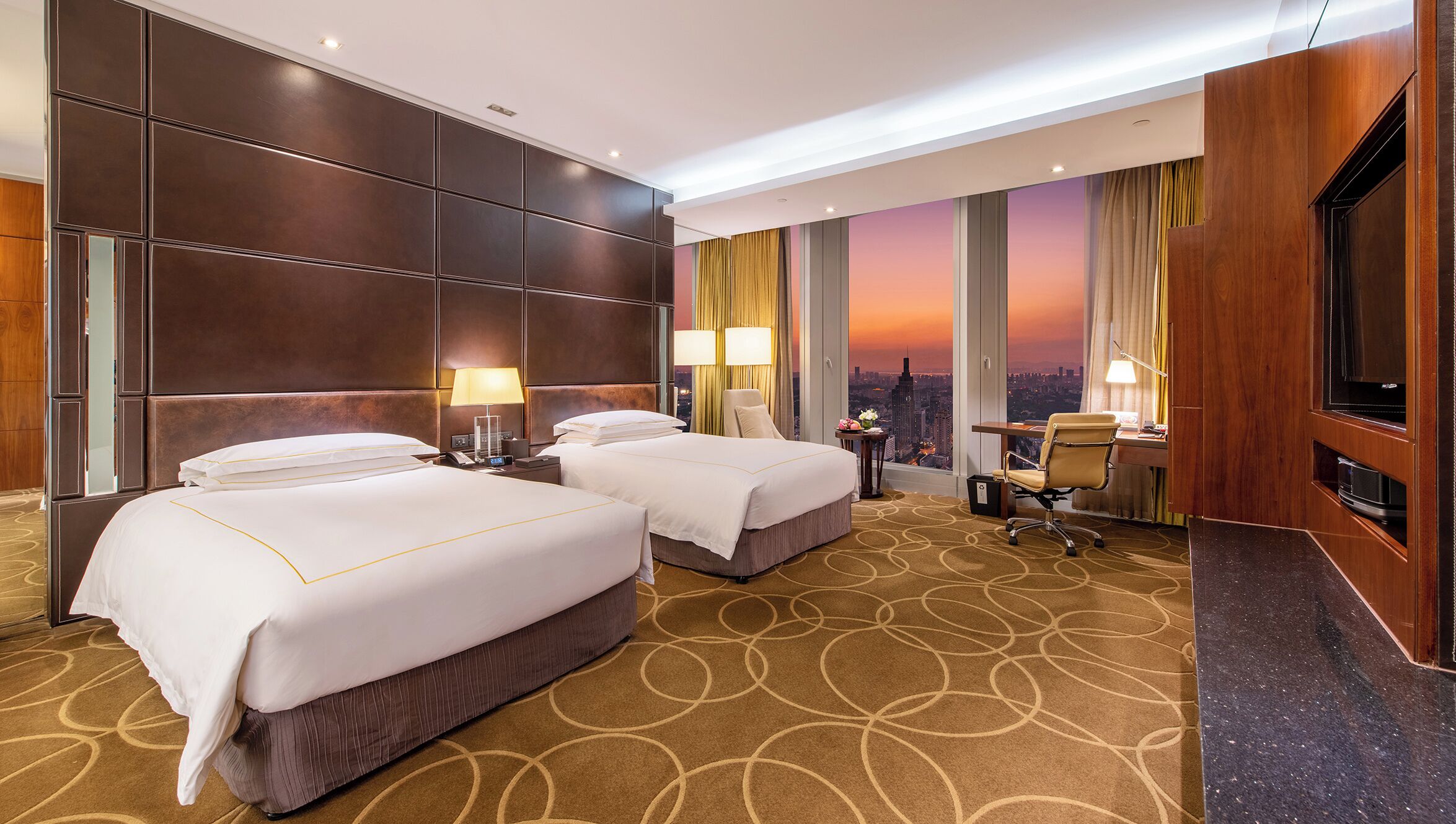 A hotel room with two beds, a desk, a chair, and a city view.