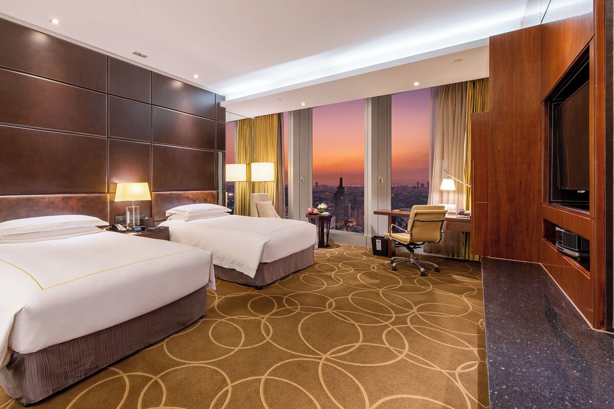 A hotel room with two beds, a desk, a chair, and a city view.