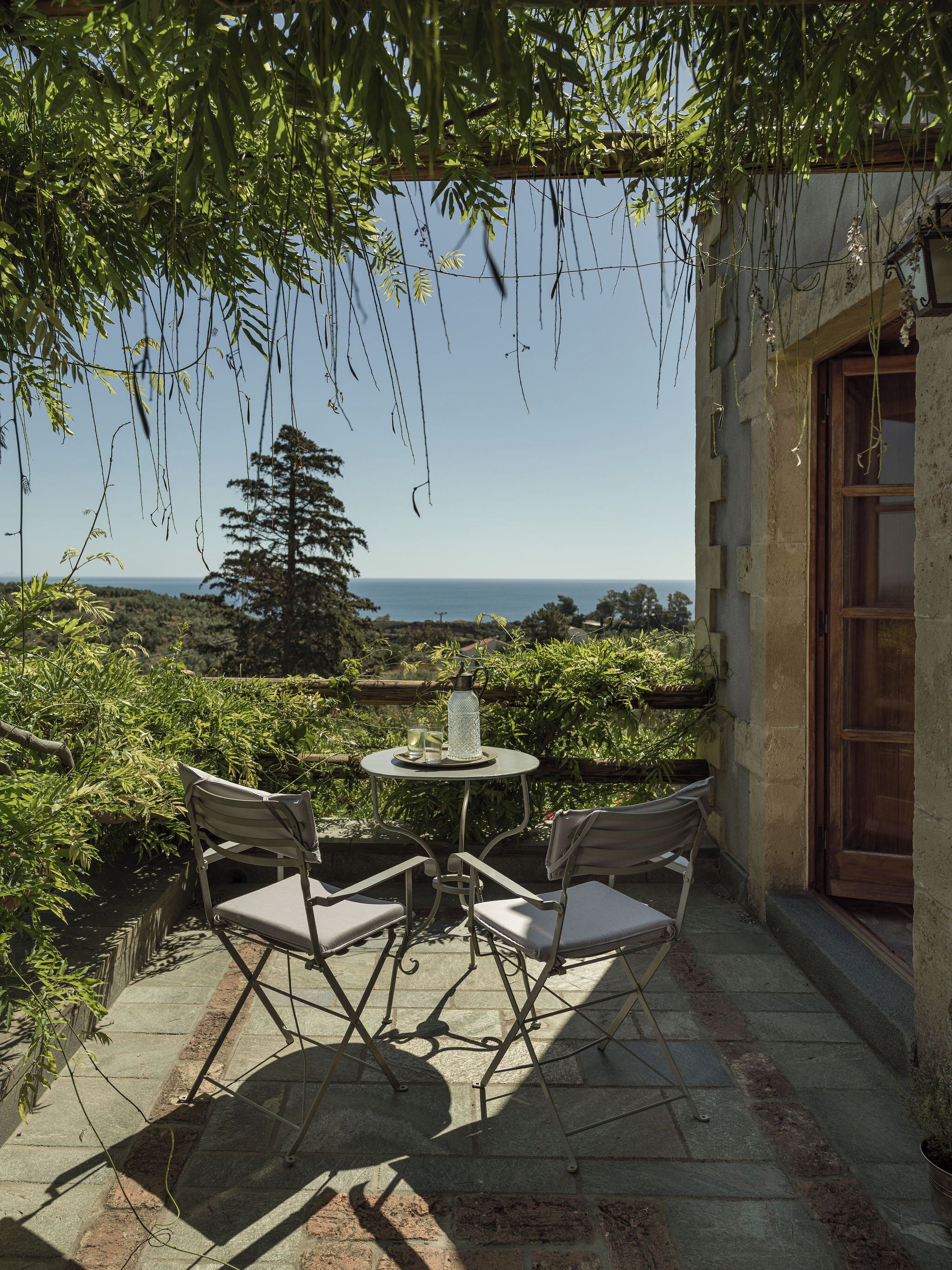 Un patio con mesa y sillas, con vista a un paisaje pintoresco y el mar, con colinas lejanas.