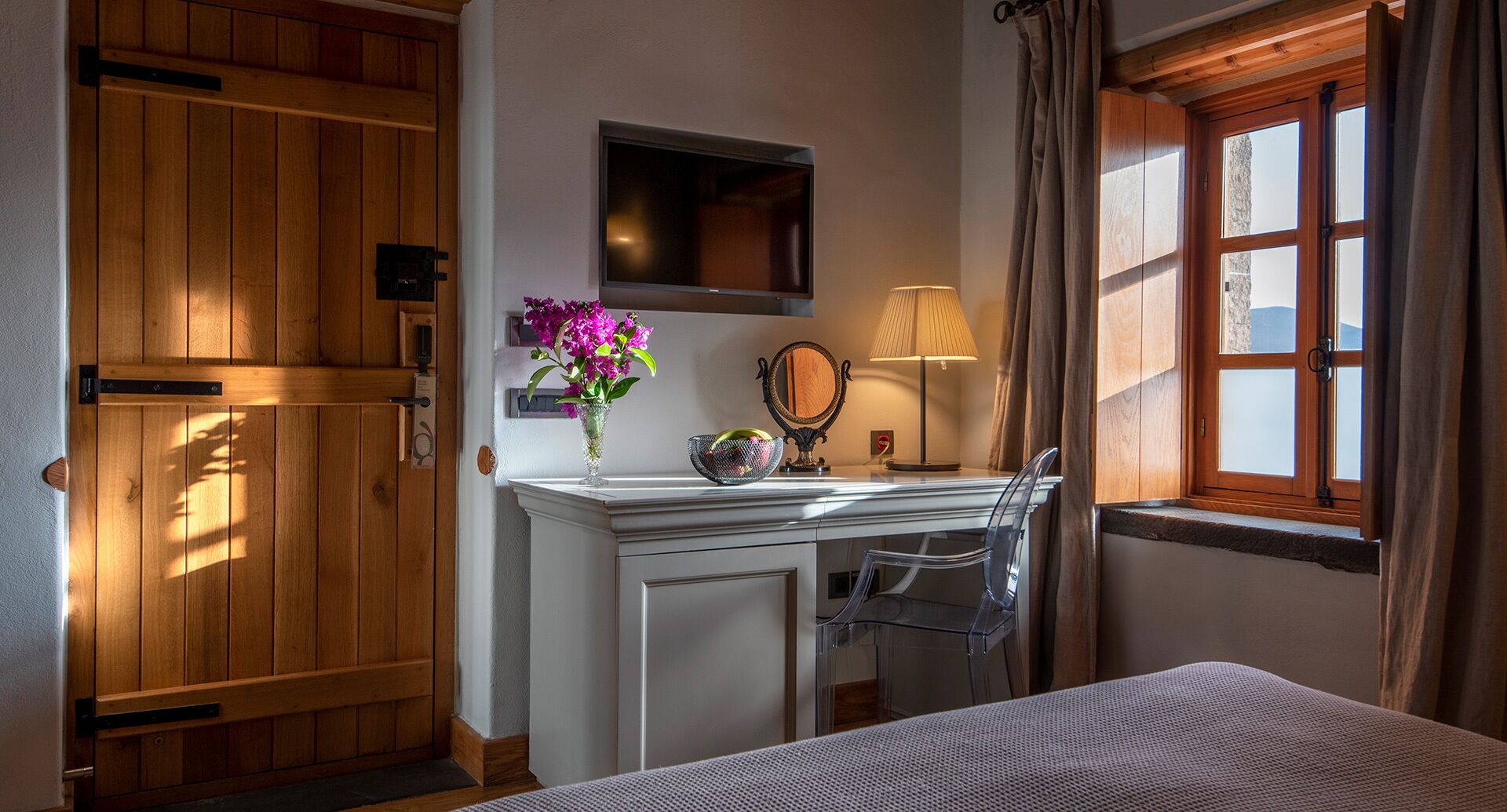 Un dormitorio con puerta de madera, una mesita de noche con lámpara, un espejo y una ventana con cortinas.