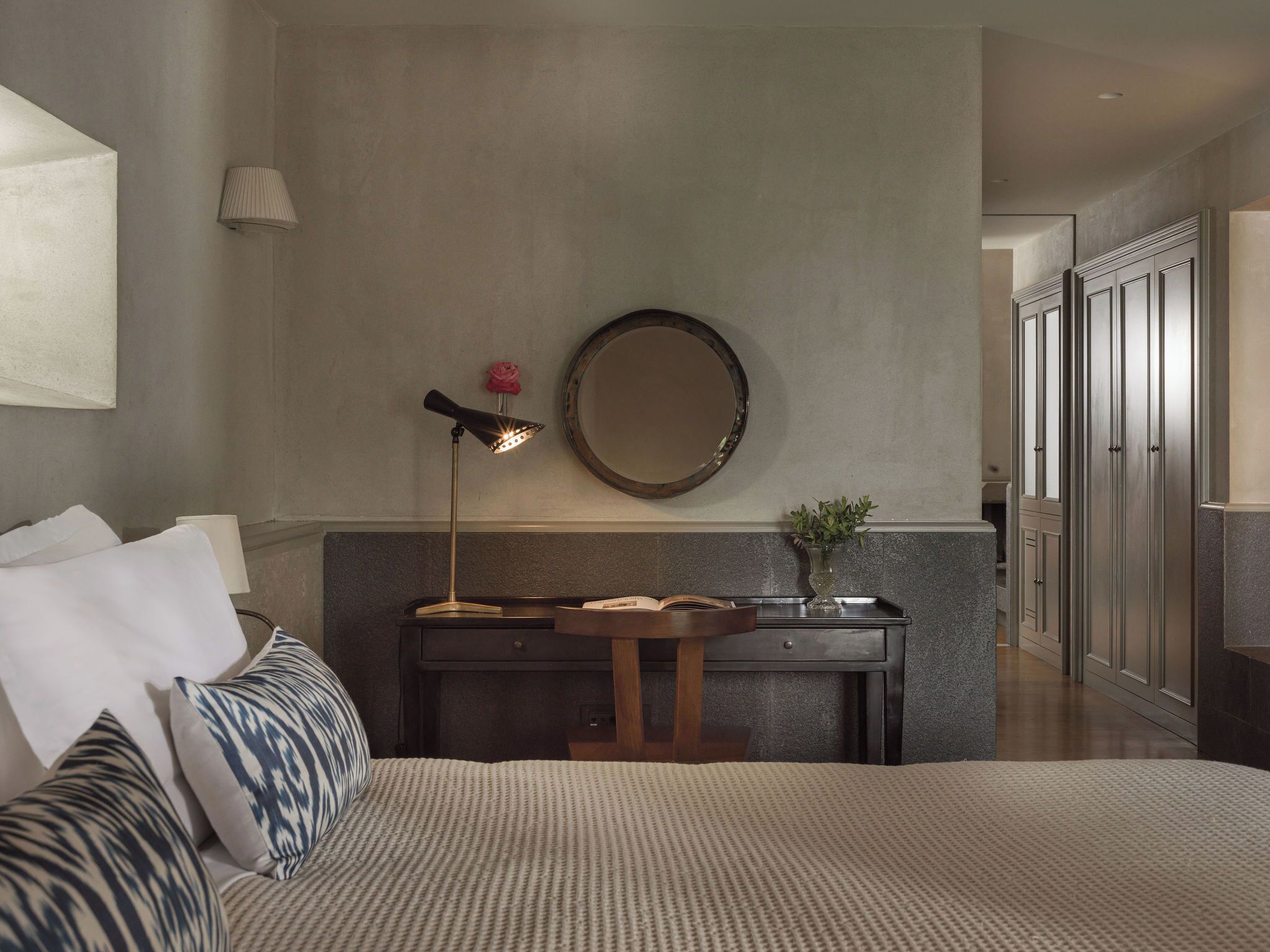 Habitación de hotel con cama, mesita de noche, lámpara, espejo y vista al pasillo.