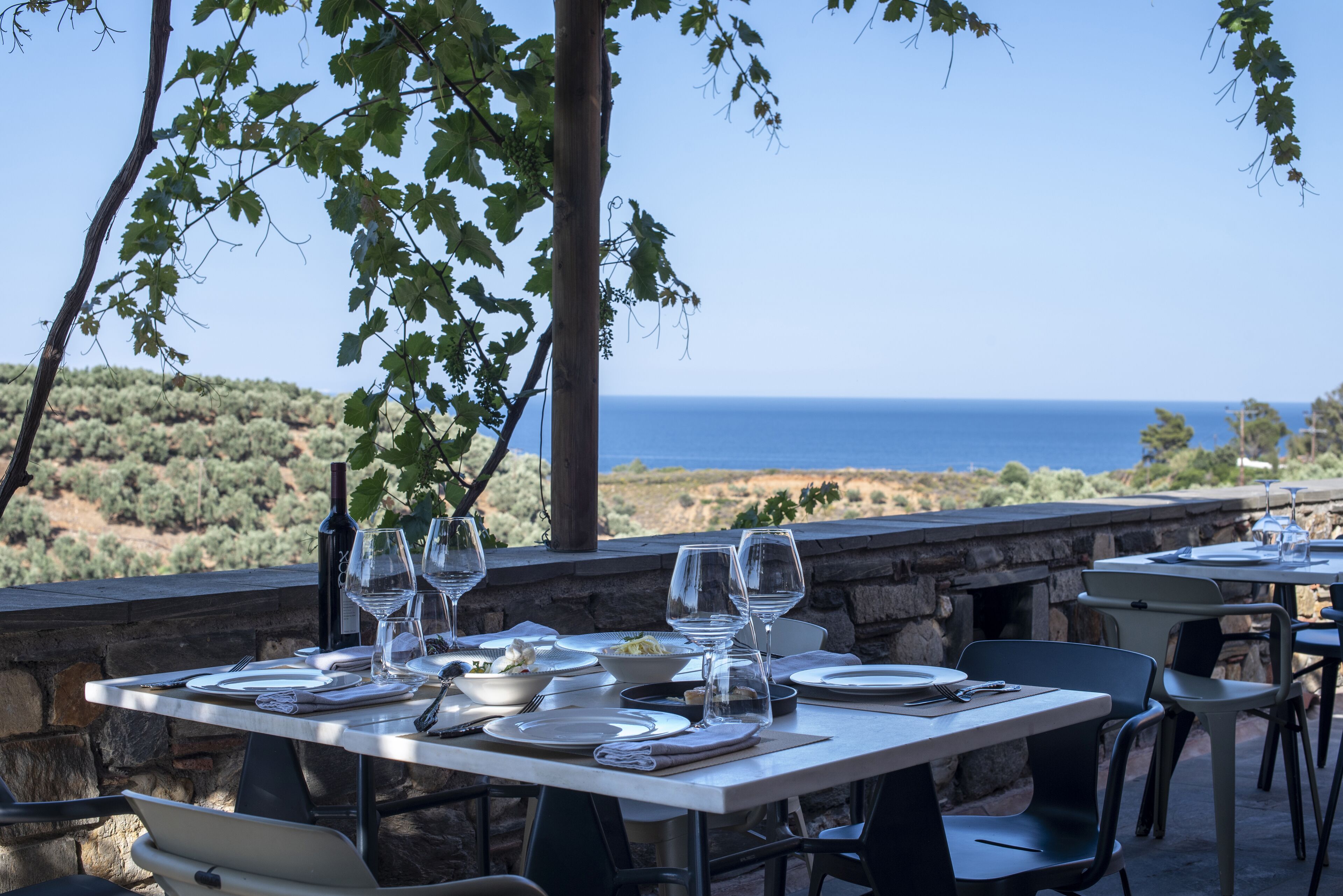 An outdoor dining area with stone walls, tables set for two, and a view of the sea.