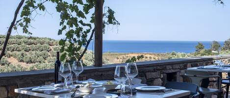 An outdoor dining area with stone walls, tables set for two, and a view of the sea.
